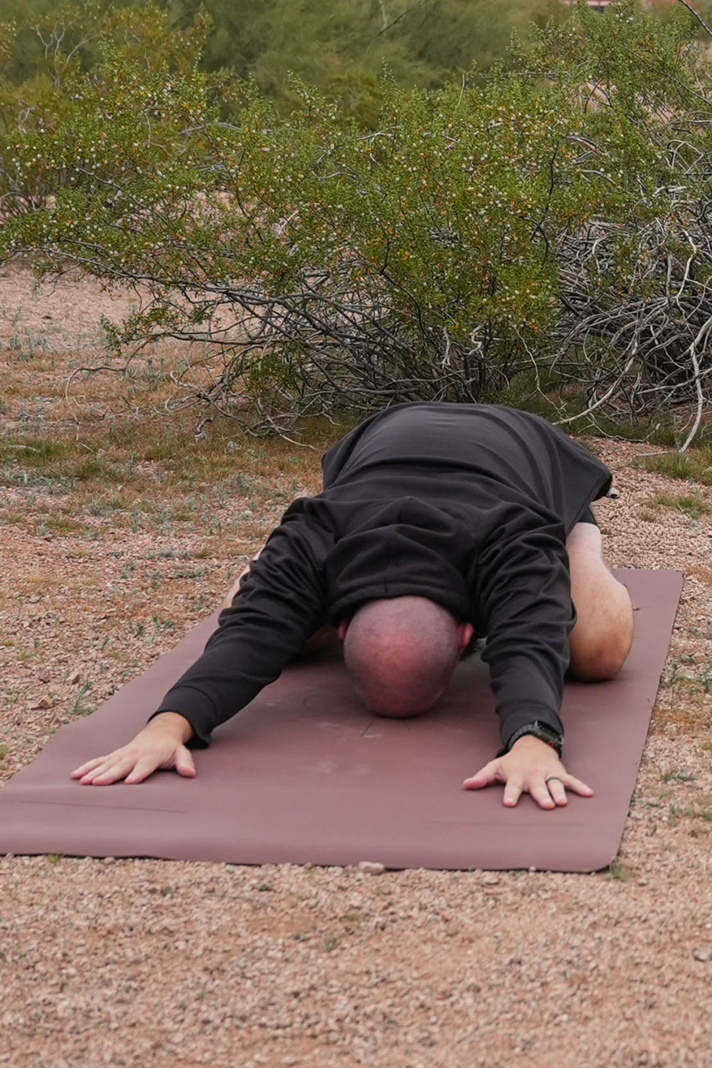 A male yoga practitioner in black long-sleeve top perform a forward bend yoga pose called Childs pose on an extra wide yoga mat in the Sedona Red Rocks color in the Sonoran Sunset Design outdoors in a sandy, brush-filled desert landscape at Papago Park, Phoenix, AZ.