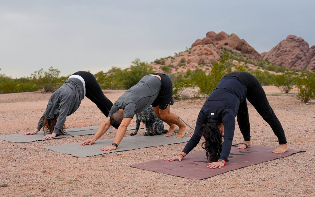 Three people in athletic wear perform a yoga pose downward dog on outdoor extra wide yoga mats in a desert landscape at Papago Park, Phoenix Arizona with rocky hills in the background.