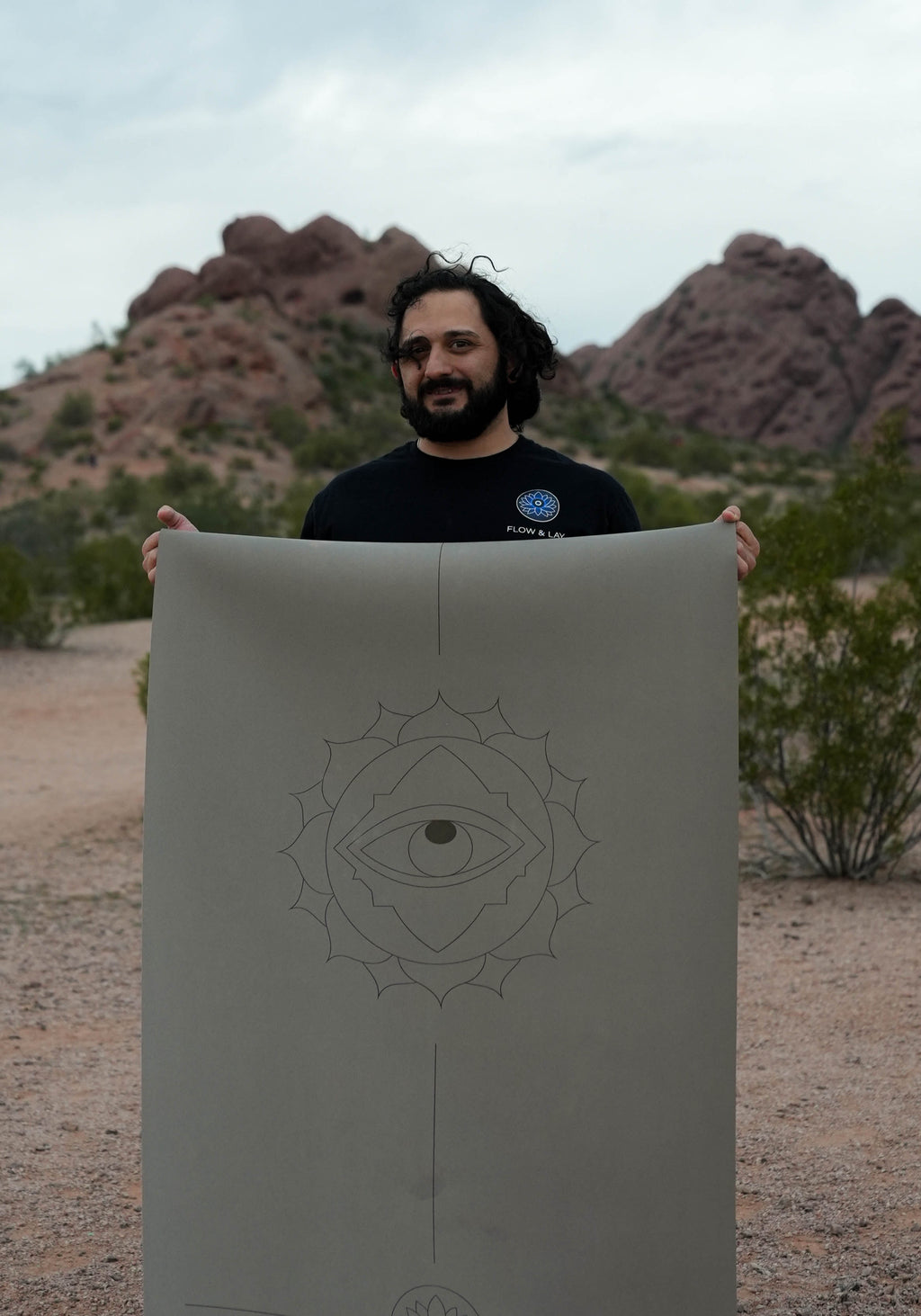 Person holding a extra tall and extra wide eclipse shadow colored yoga mat with the throat eye design in the Papago Park desert in Phoenix Arizona.