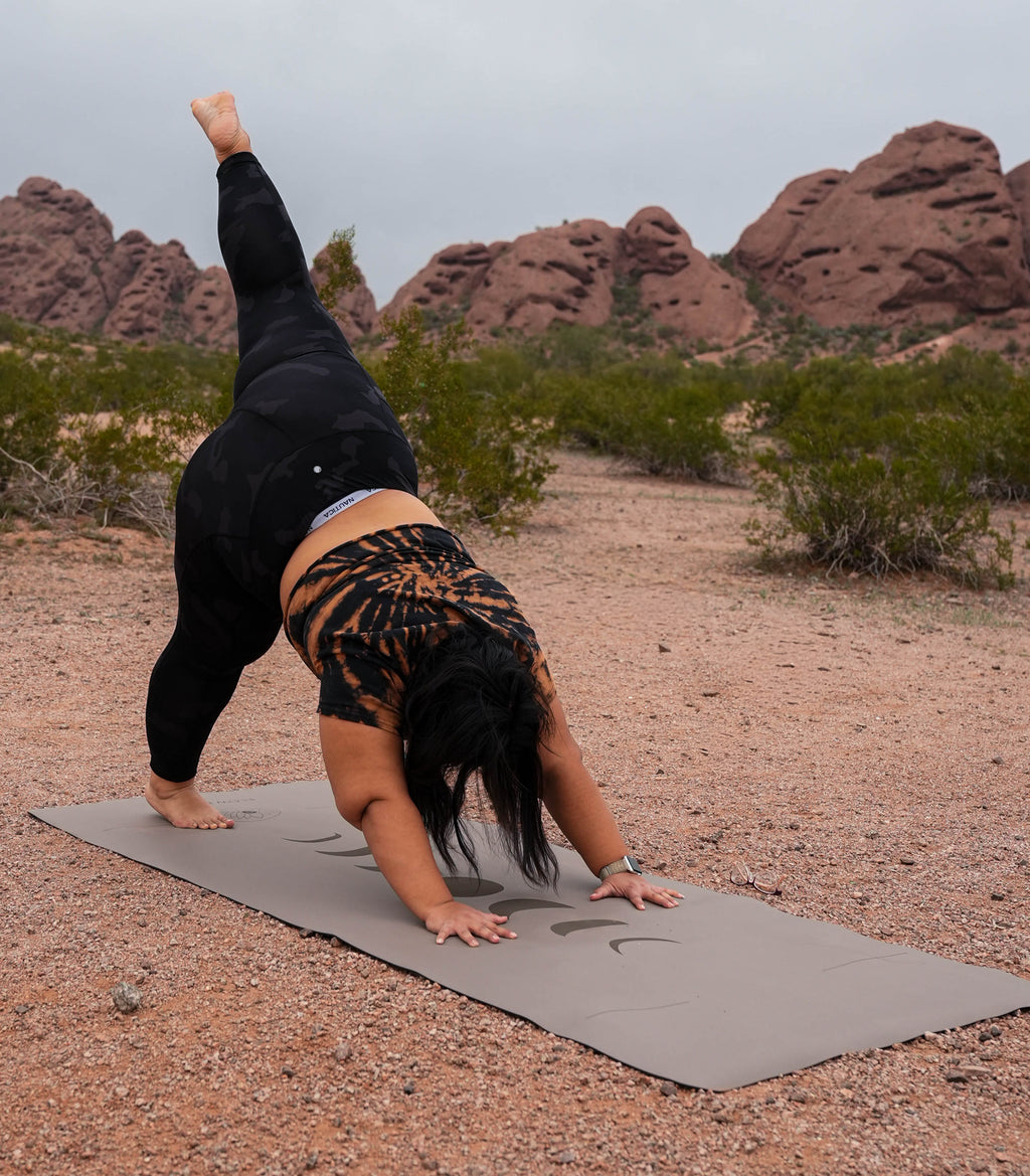 A woman in a black activewear set performs a yoga pose downward dog on the Eclipse Shadow Extra Wide and Extra Tall yoga mat in a desert landscape at Papago Park Phoenix, Arizona with red rock formations in the background. The design is Phases of the moon.