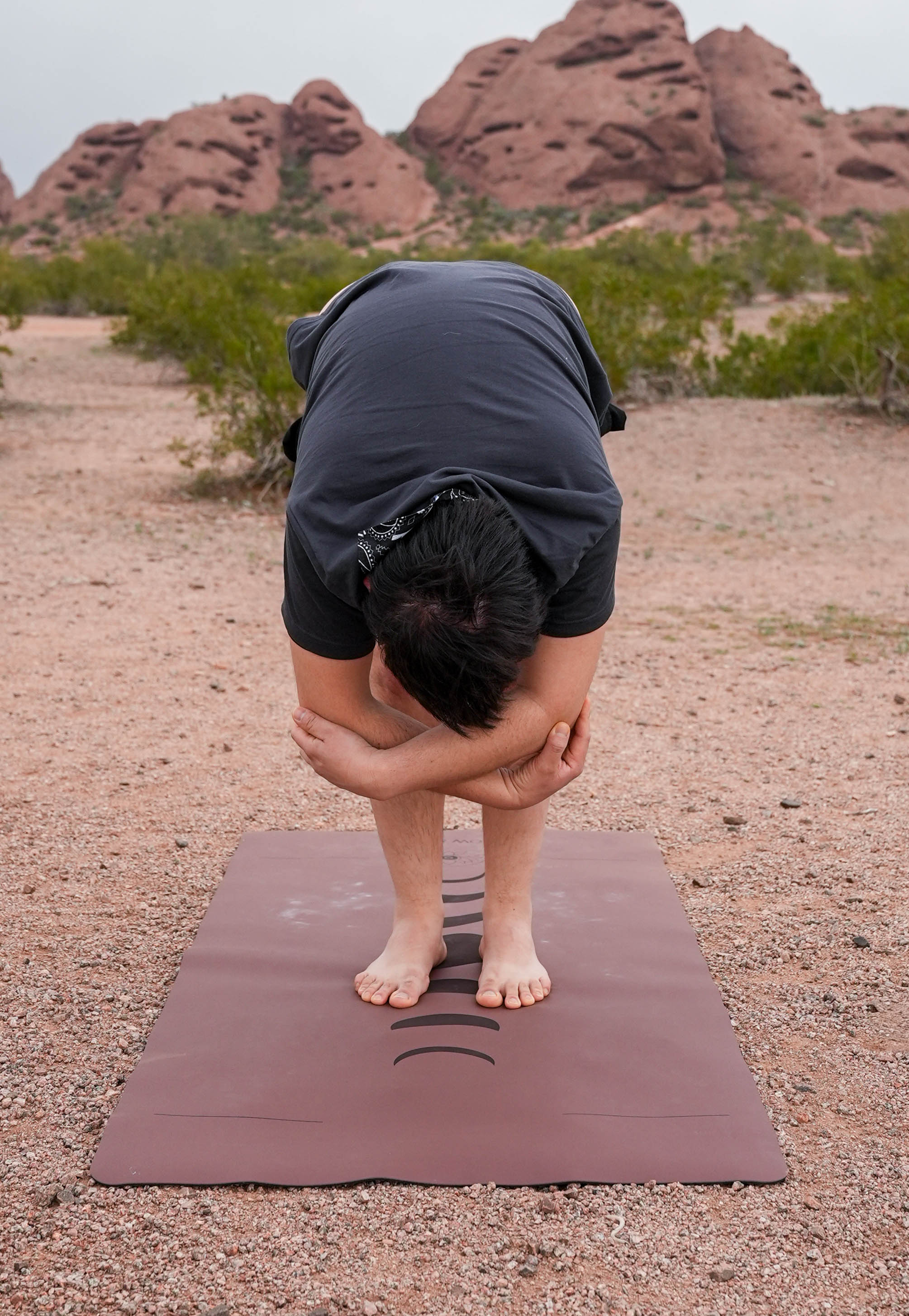 A man performing a forward bend yoga pose on a Red Rocks Sedona yoga mat, with a desert landscape of red rock formations in Papago Park Phoenix Arizona and green shrubs in the background.