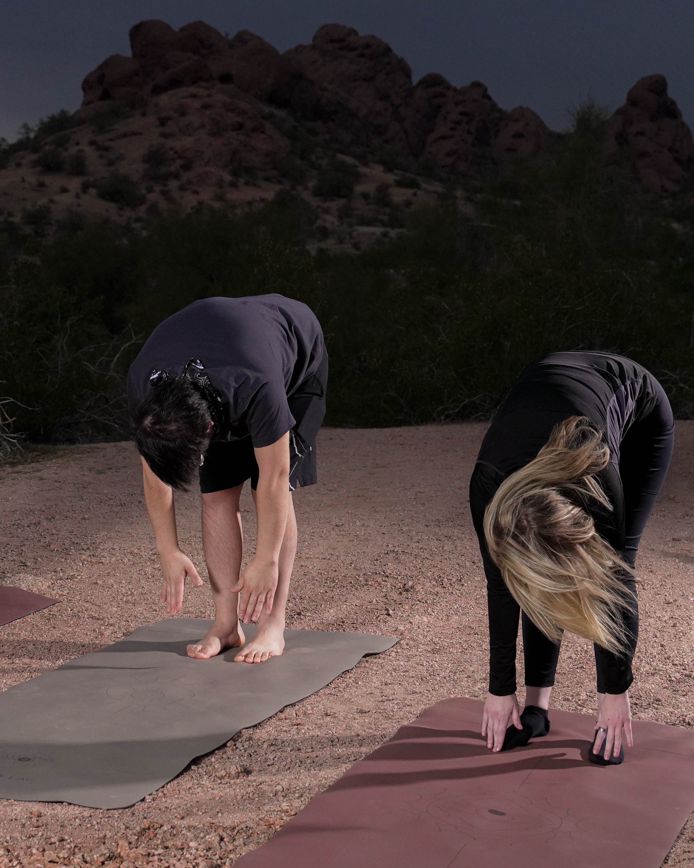 Two women practice yoga outdoors at Papago Park in Phoenix  on a sandy desert path at night, with rocky mountains and dark sky in the background. They are positioned on Flow & Lay extra wide yoga mats: an eclipse shadow colored mat in the center, and a red rocks Sedona colored mat on the right.