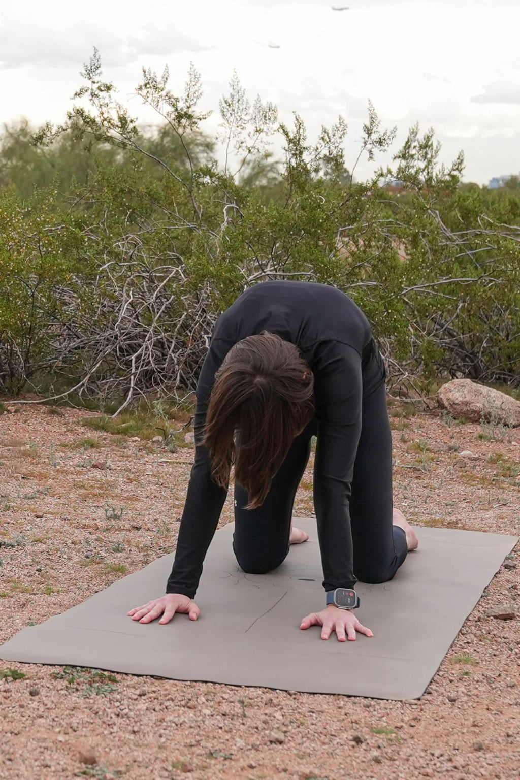 Woman in black activewear protecting their knees using extra wide yoga mats by Flow & Lay while doing a cat-cow stretch outdoors on a desert sandy ground with shrubs and rocks in the background.