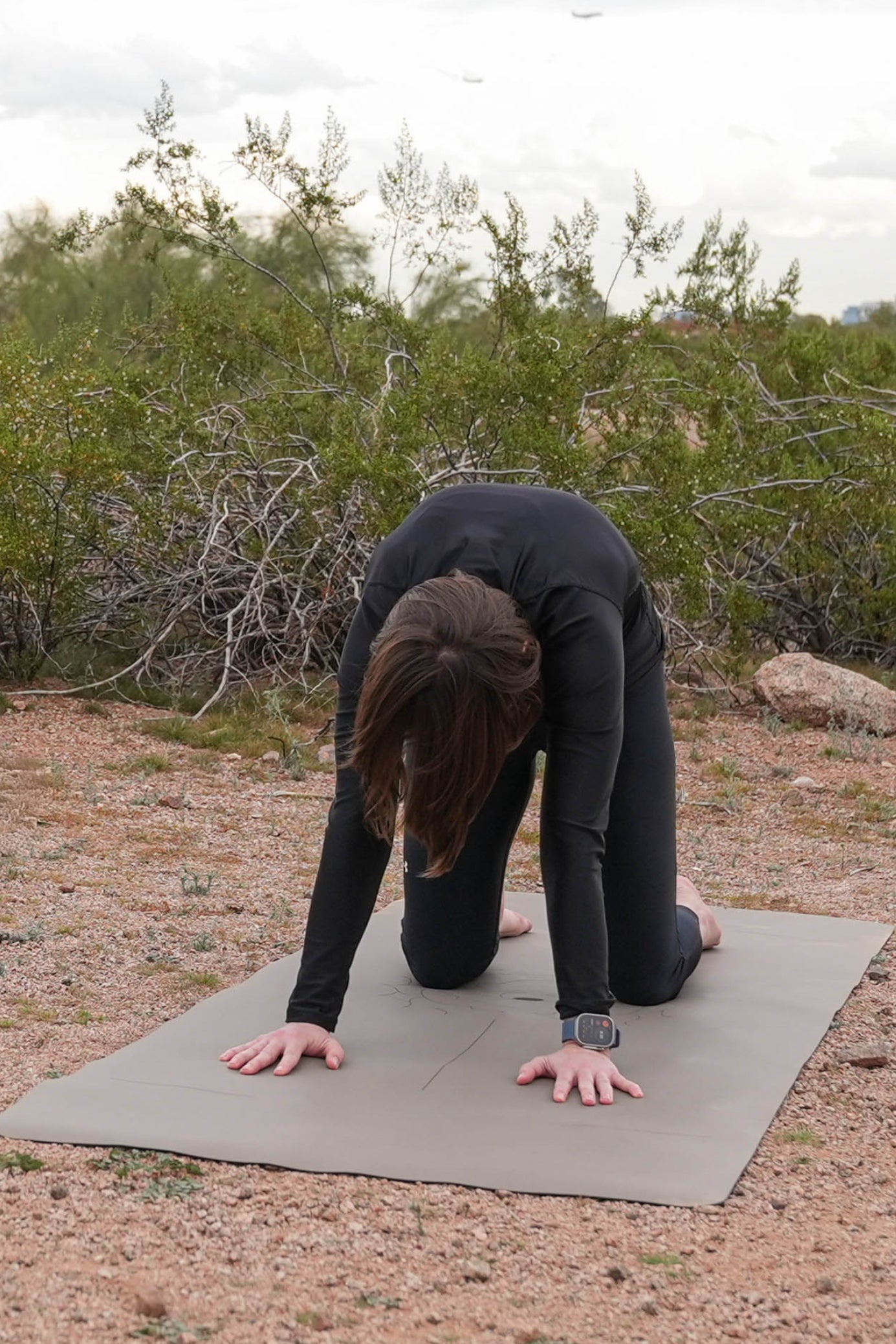 Woman in black activewear protecting their knees using extra wide yoga mats by Flow & Lay while doing a cat-cow stretch outdoors on a desert sandy ground with shrubs and rocks in the background.