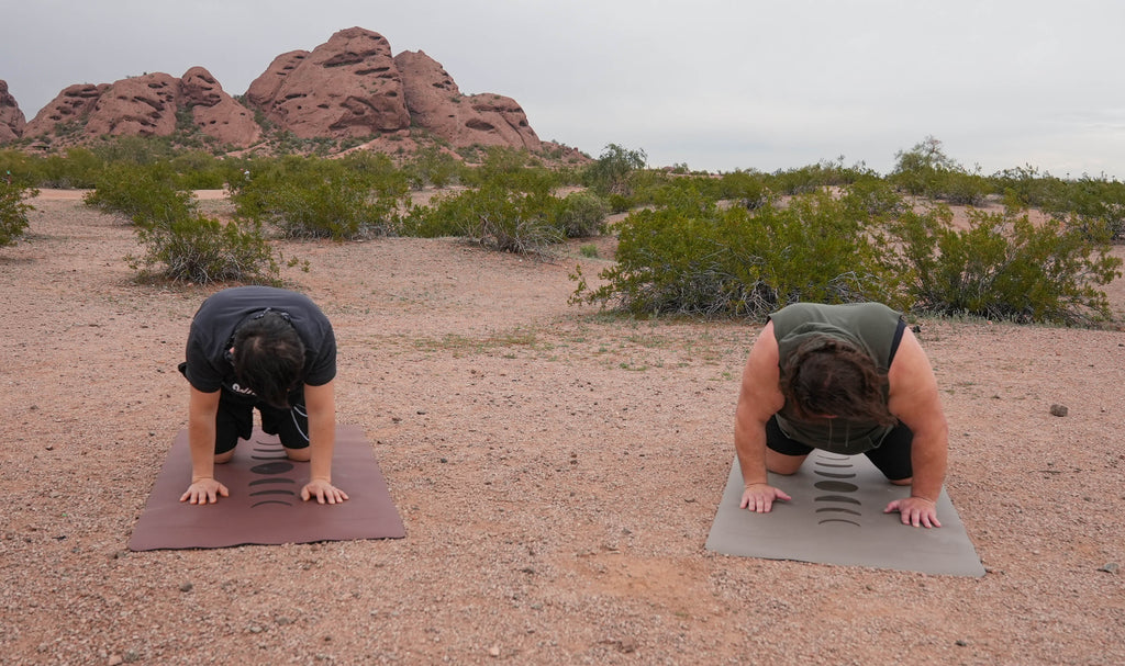 Two people in desert terrain performing push-up exercises on extra wide Phases of the Moon yoga mats, with red rock formations at Papago Park, Phoenix Arizona and shrubs in the background.