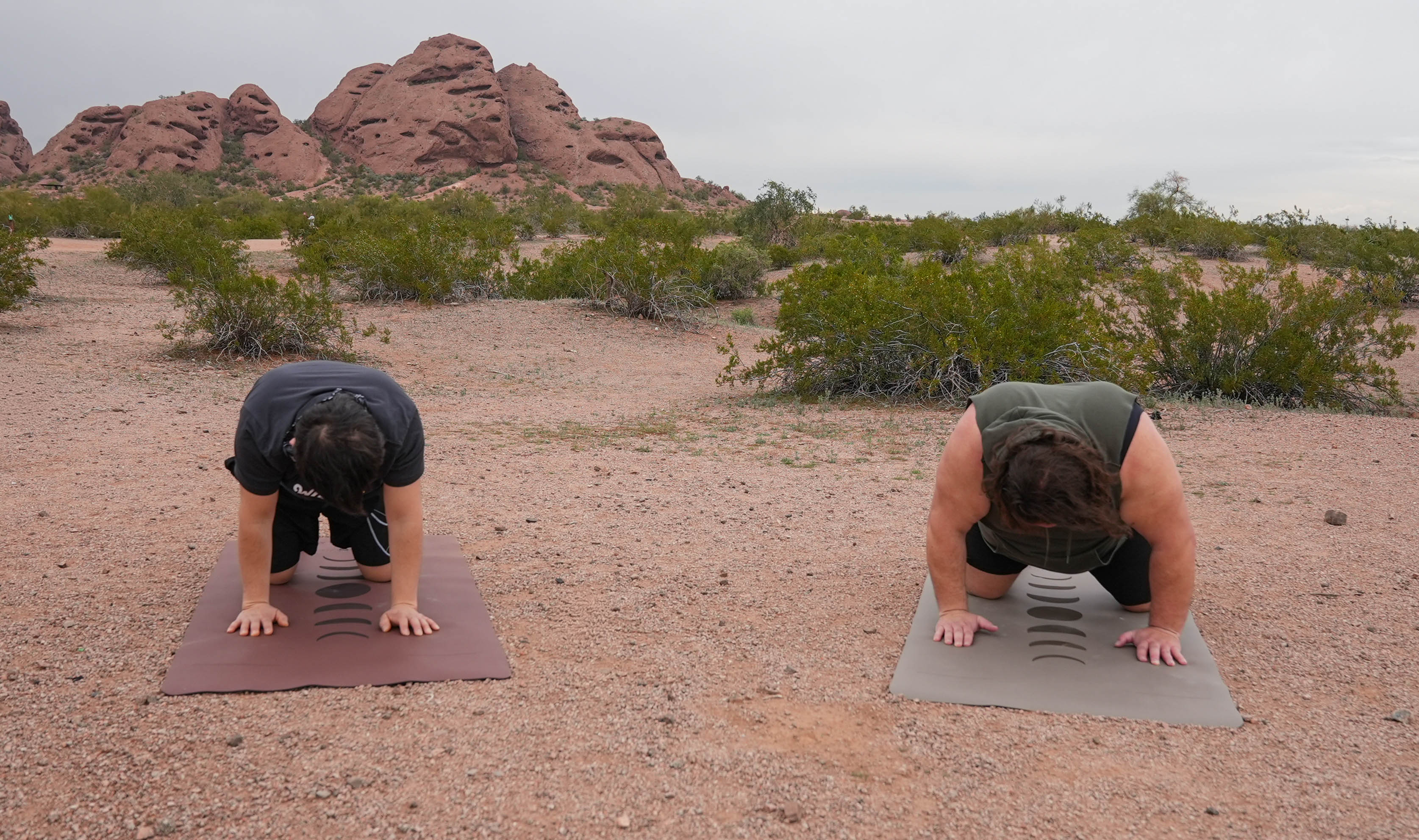 Two people in desert terrain performing push-up exercises on extra wide Phases of the Moon yoga mats, with red rock formations at Papago Park, Phoenix Arizona and shrubs in the background.