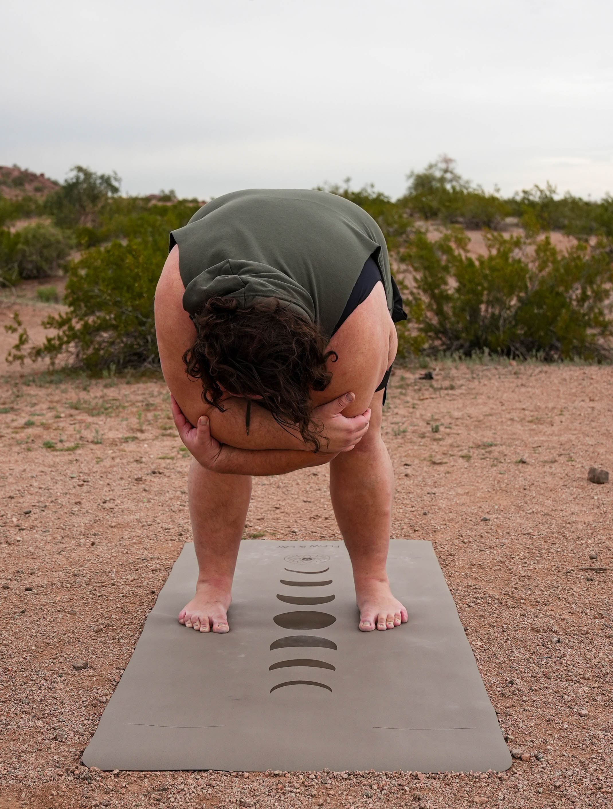A person in an outdoor desert setting in Papago Park,Phoenix Arizona performs a deep forward bend on an extra wide extra tall yoga mat printed with phases of the moon, wearing an olive green top and black shorts.