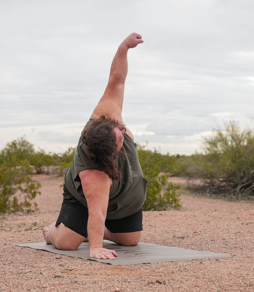 A person outdoors in a desert setting, kneeling on a yoga mat and stretching their body into a side pose with one arm reaching upward. They are wearing an olive green sleeveless top and black shorts. Phases of the moon extra wide extra tall yoga mat in eclipse shadow color.