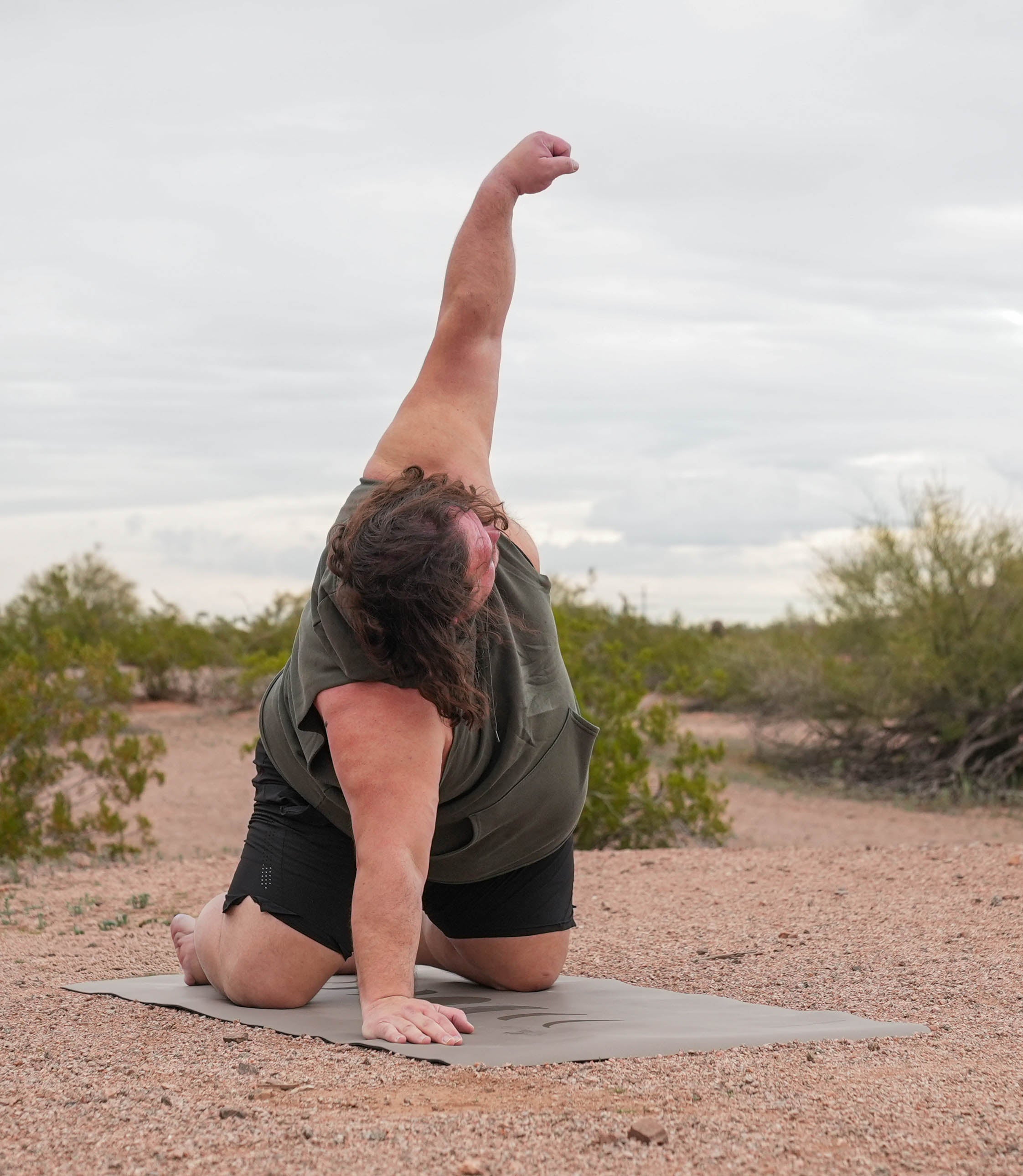 A person outdoors in a desert setting, kneeling on a yoga mat and stretching their body into a side pose with one arm reaching upward. They are wearing an olive green sleeveless top and black shorts. Phases of the moon extra wide extra tall yoga mat in eclipse shadow color.