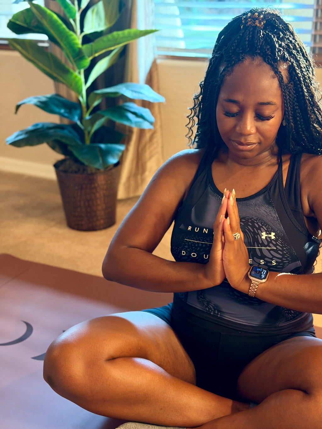 A pregnant woman sits cross-legged on an extra wide yoga mat in an Arizona home setting, wearing a black athletic tank top with a graphic print and black shorts. She is in a meditative pose with her hands together in Anjali Mudra , with a potted plant and window in the background.