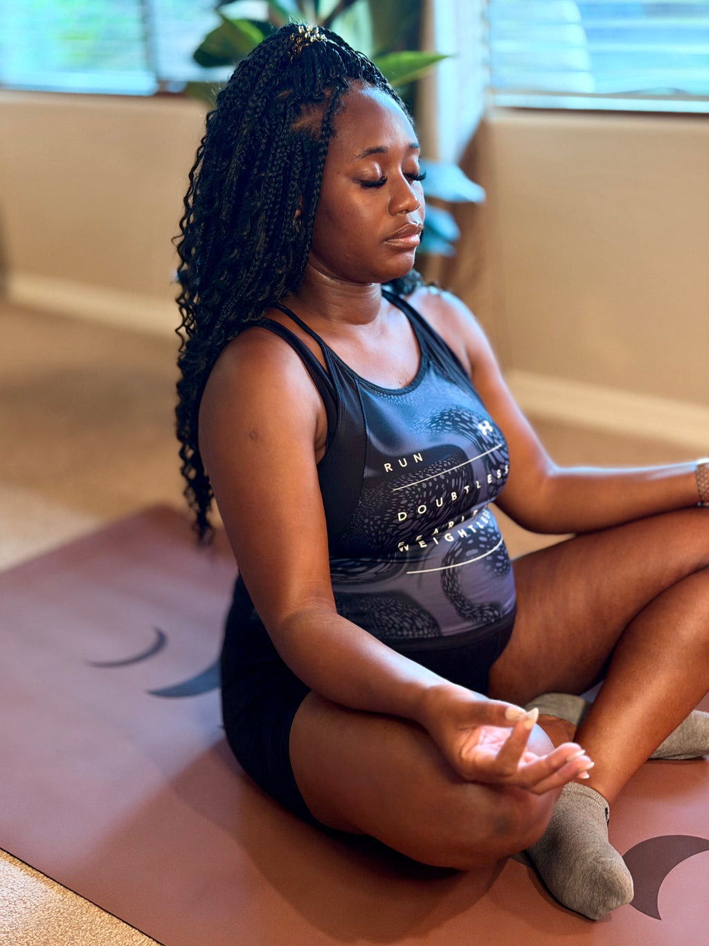 A pregnant woman meditating in a yoga pose on an extra wide yoga mat in Red Rocks Sedona color with Phases of the moon design, wearing a black tank top with a gray graphic print and black shorts in a brightly lit room in Phoenix, Arizona.