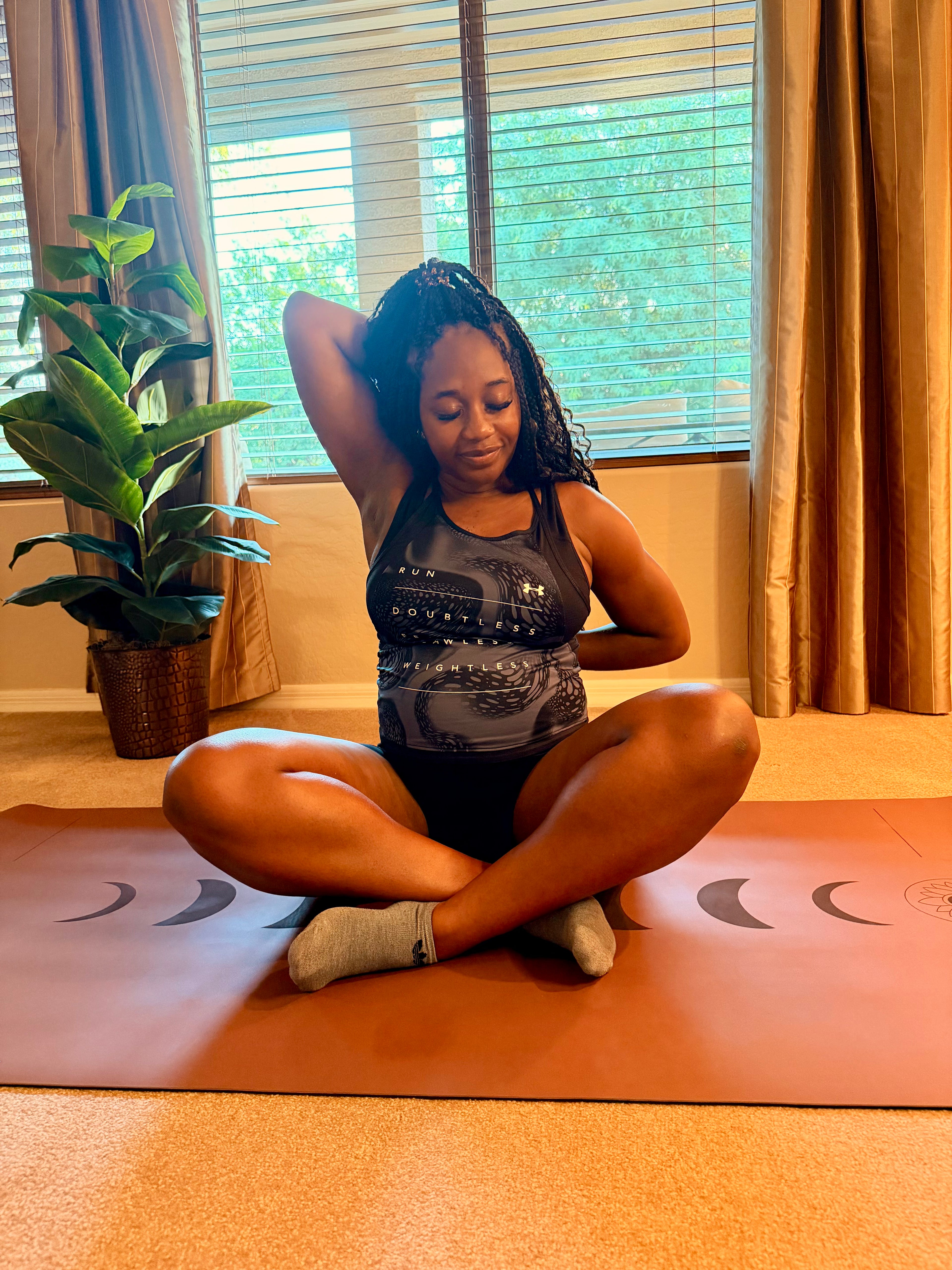 A pregnant woman sits cross-legged on an extra wide yoga mat Red Rocks Sedona Color in Phases of the moon design in an Arizona home setting, wearing a gray patterned tank top and black shorts, stretching her arms overhead called gomukhasana arms near a window with curtains and a potted plant.