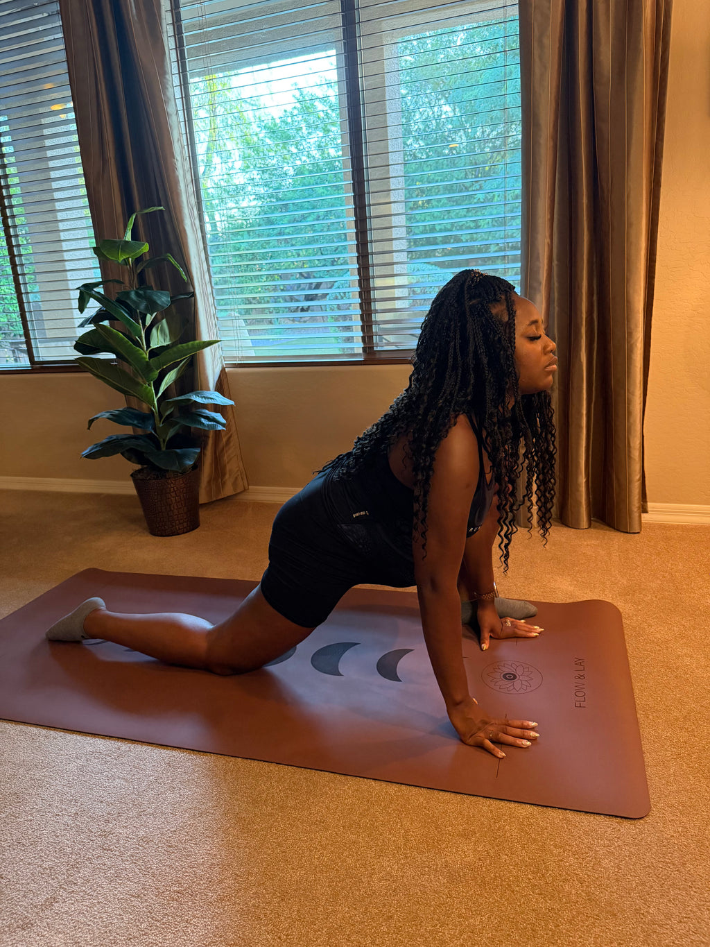 A pregnant woman in a black athletic outfit performs a yoga pose for a deep hip stretch, lizard lunge, on an extra wide yoga mat in an Arizona home setting with beige carpet, windows with blinds, and a potted plant.