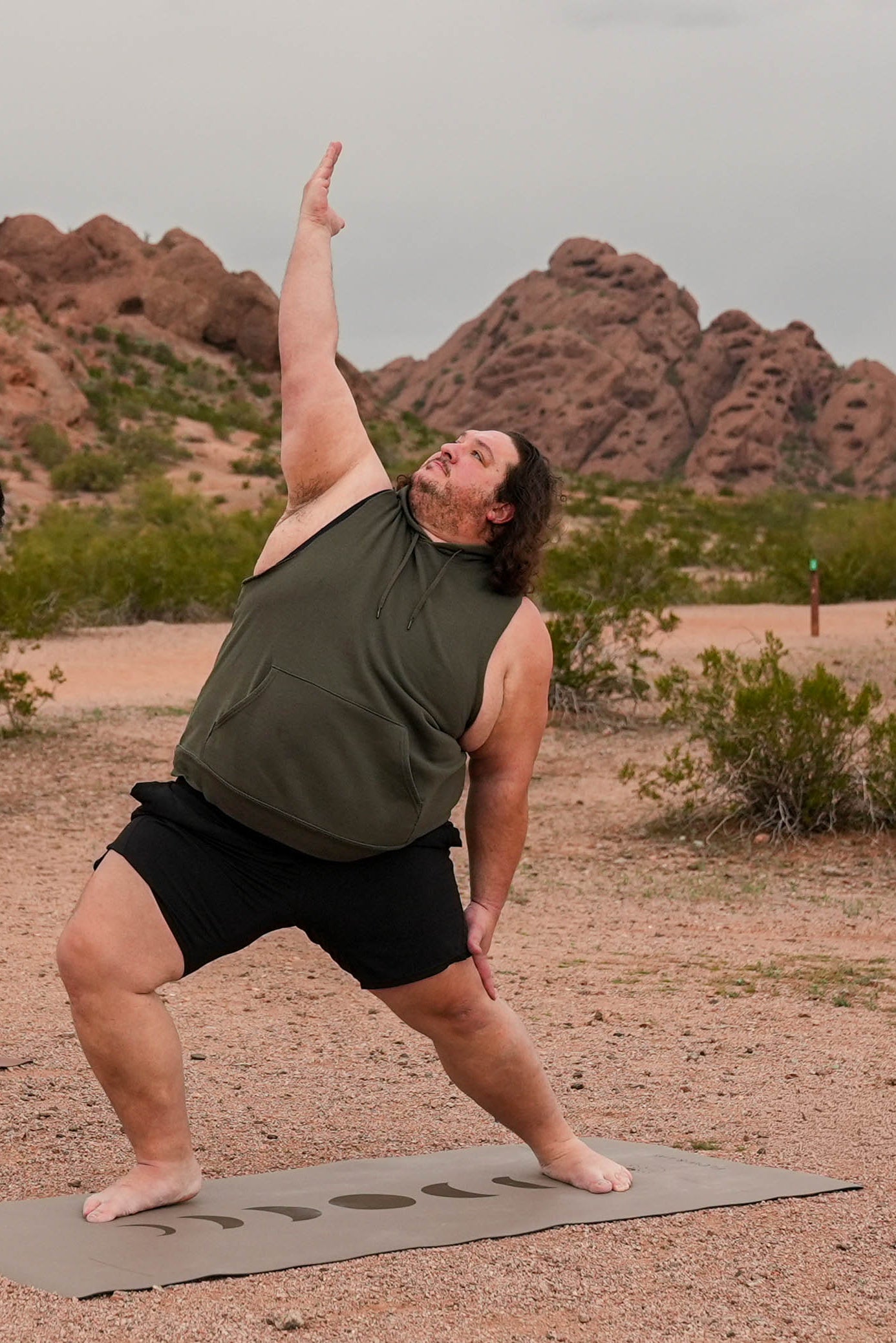 Man performing yoga pose reverse warrior outdoors in a desert landscape with red rock formations in the background at Papago Park, Phoenix Arizona. The man in the foreground wears an olive green tank top, black shorts, and an eclipse shadow colored yoga mat designed with phases of the moon on an extra wide extra tall sized mat. 