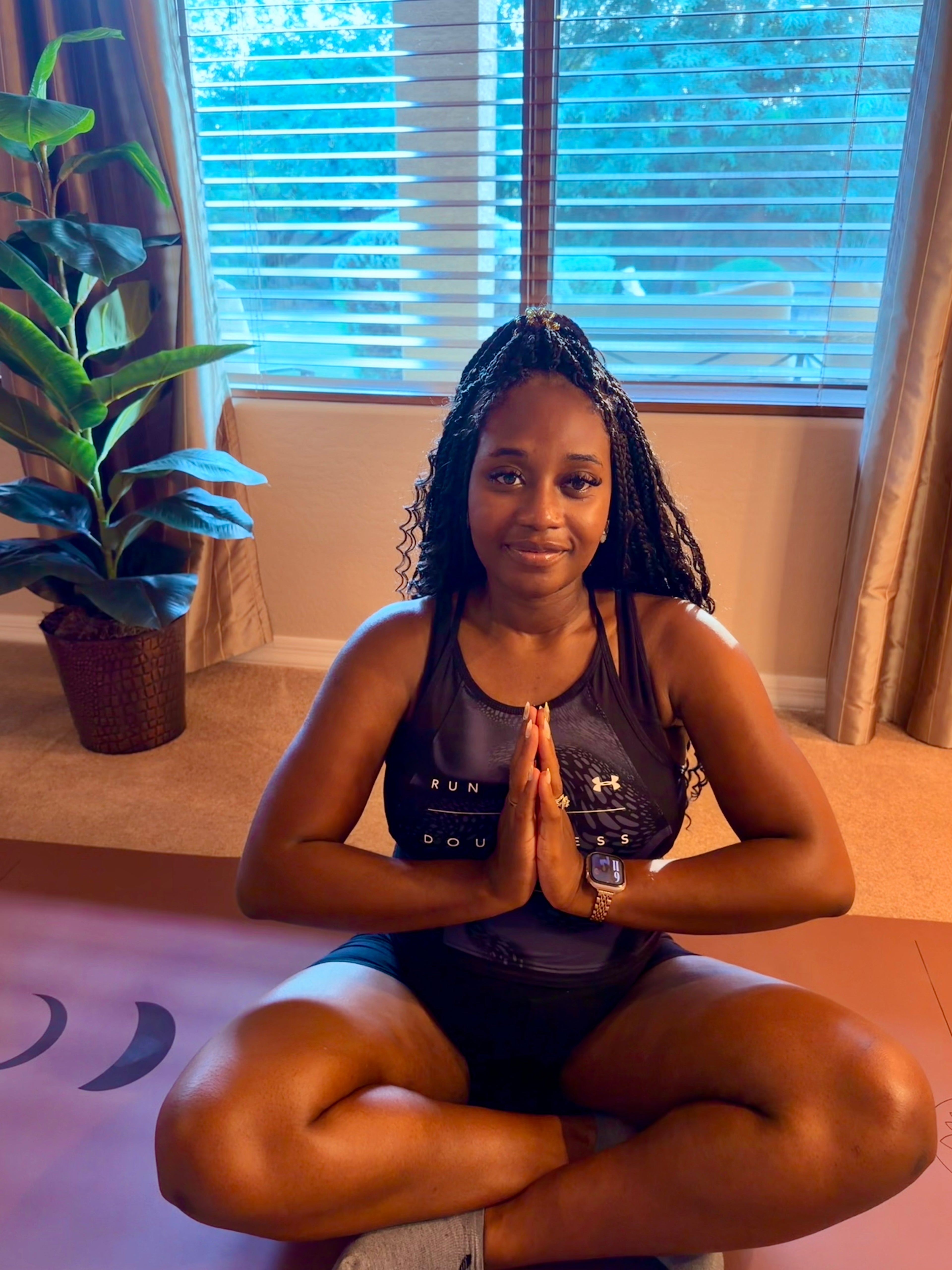 A pregnant woman sits cross-legged on an extra wide yoga mat in the Red Rocks Sedona color on the Phases of the moon design indoors, wearing a dark athletic tank top and shorts. The tank top has 'RUN SOUVENIR' and an Under Armour logo visible. Anjali Sudra hands in sukasana. 
