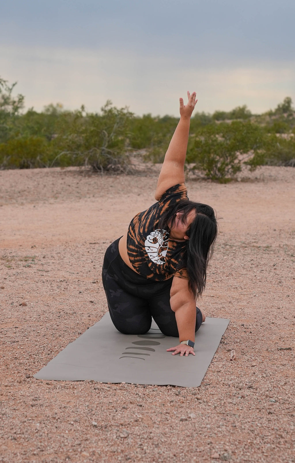 A woman in thread the needle stretch yoga pose on an eclipse shadow Phases of the Moon extra wide yoga mat in an outdoor desert setting at Papago Park, Phoenix Arizona, wearing a tiger-print crop top and black leggings.