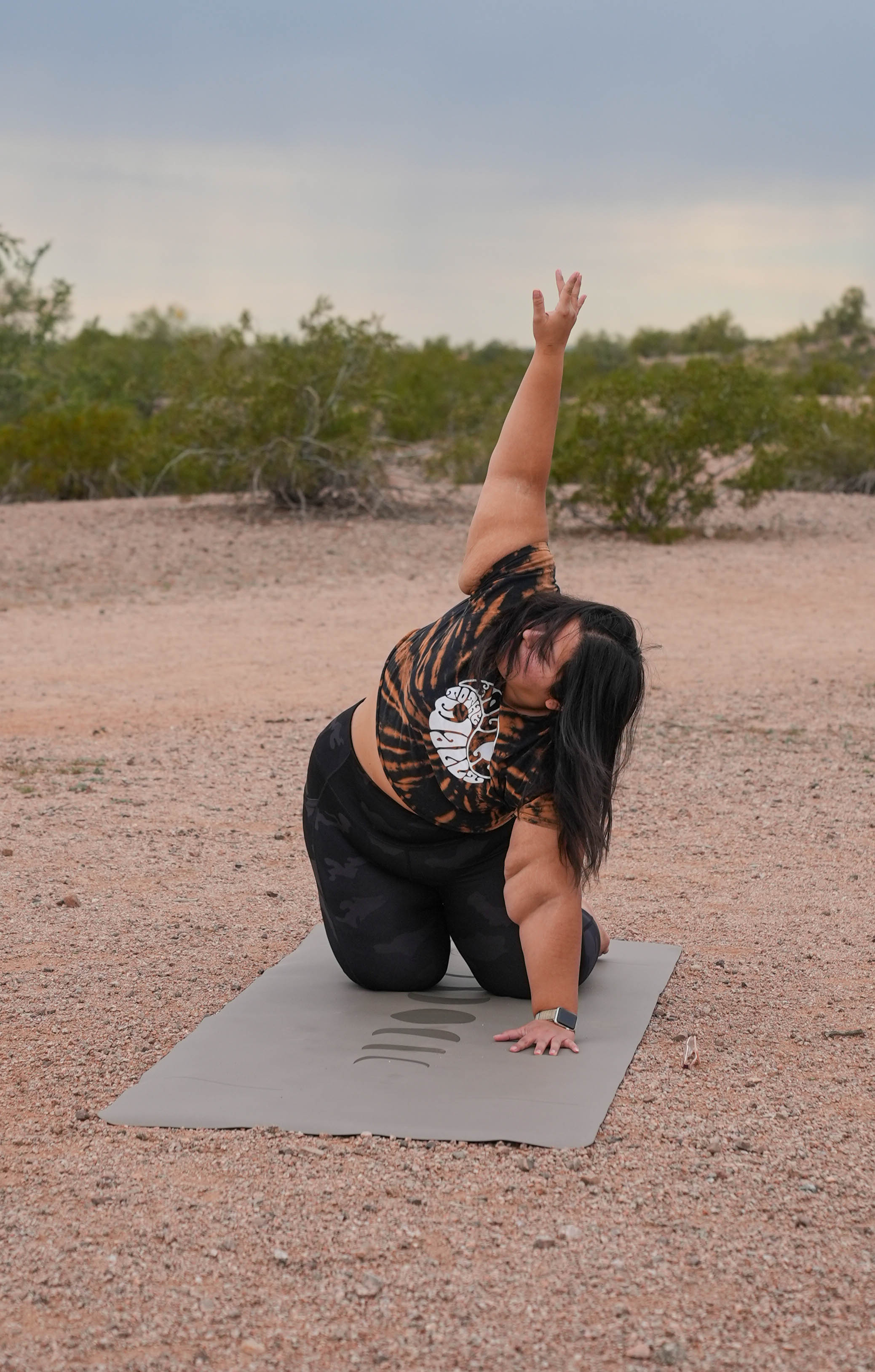 A woman in thread the needle stretch yoga pose on an eclipse shadow Phases of the Moon extra wide yoga mat in an outdoor desert setting at Papago Park, Phoenix Arizona, wearing a tiger-print crop top and black leggings.
