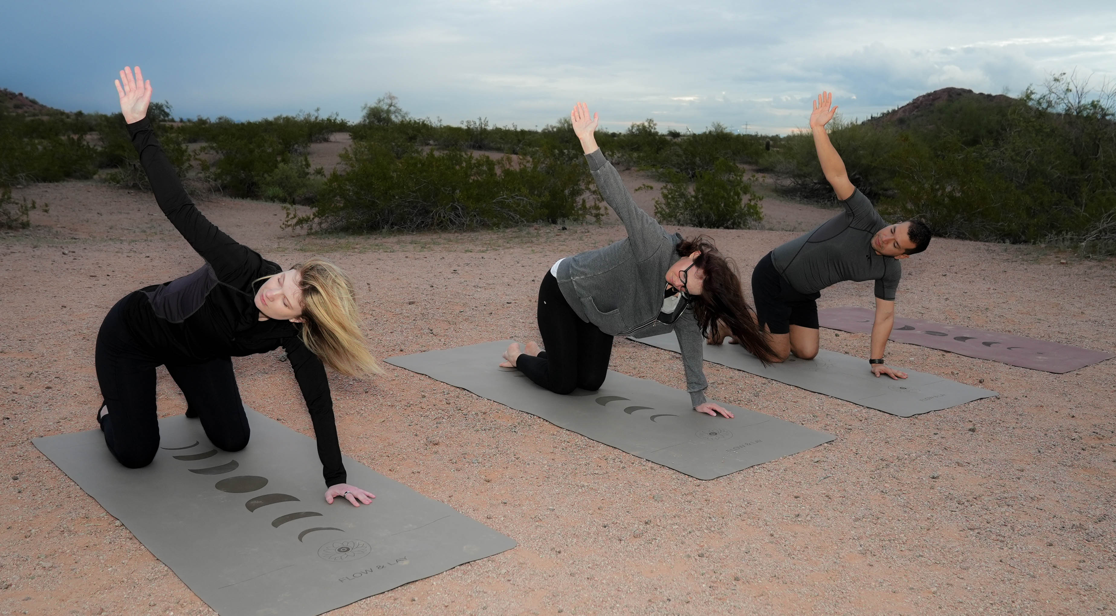 Three people practicing yoga outdoors in a desert setting at Papago Park, Phoenix Arizona, performing thread the needle pose on eclipse shadow yoga mats.