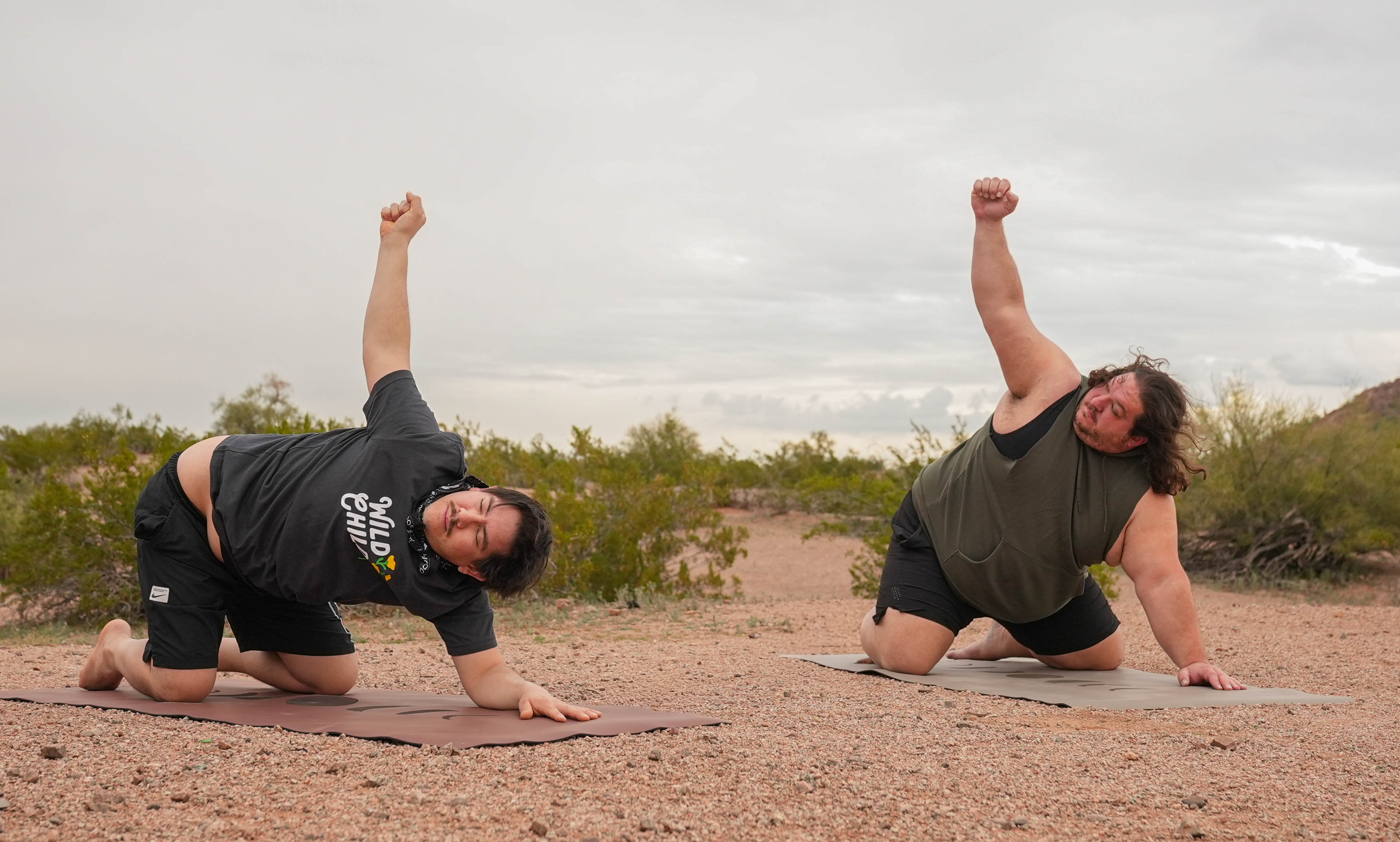 Two men performing thread the needle yoga pose outdoors at Papago Park Phoenix Arizona on a gravel path under an overcast sky, with shrubs in the background.