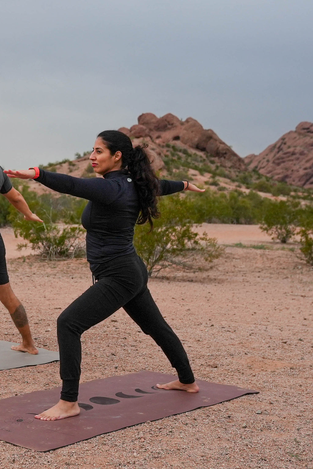 A woman in a black activewear set performs a yoga warrior 2 pose on a Red Rocks Sedona colored yoga mat in an outdoor desert landscape with red rock formations under a cloudy sky in Papago Park, Phoenix Arizona.