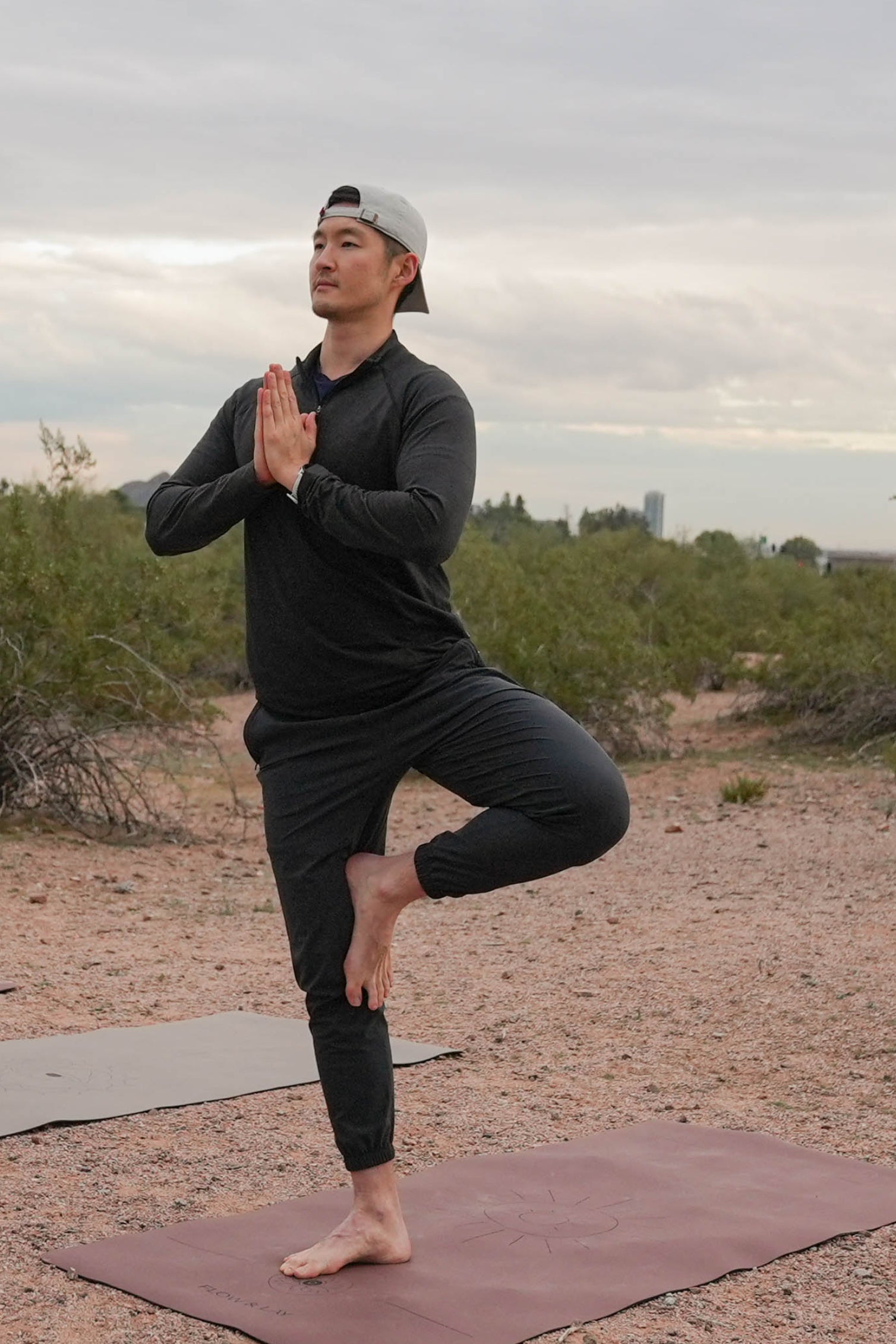 A tall man in black activewear perform tree pose yoga outdoors on a gravel path with shrubs and a cloudy sky in the background at Papago Park, Phoenix, AZ. He is doing yoga on an extra wide yoga mat in the Sedona Red Rocks Color with a Sonoran Sunset Design